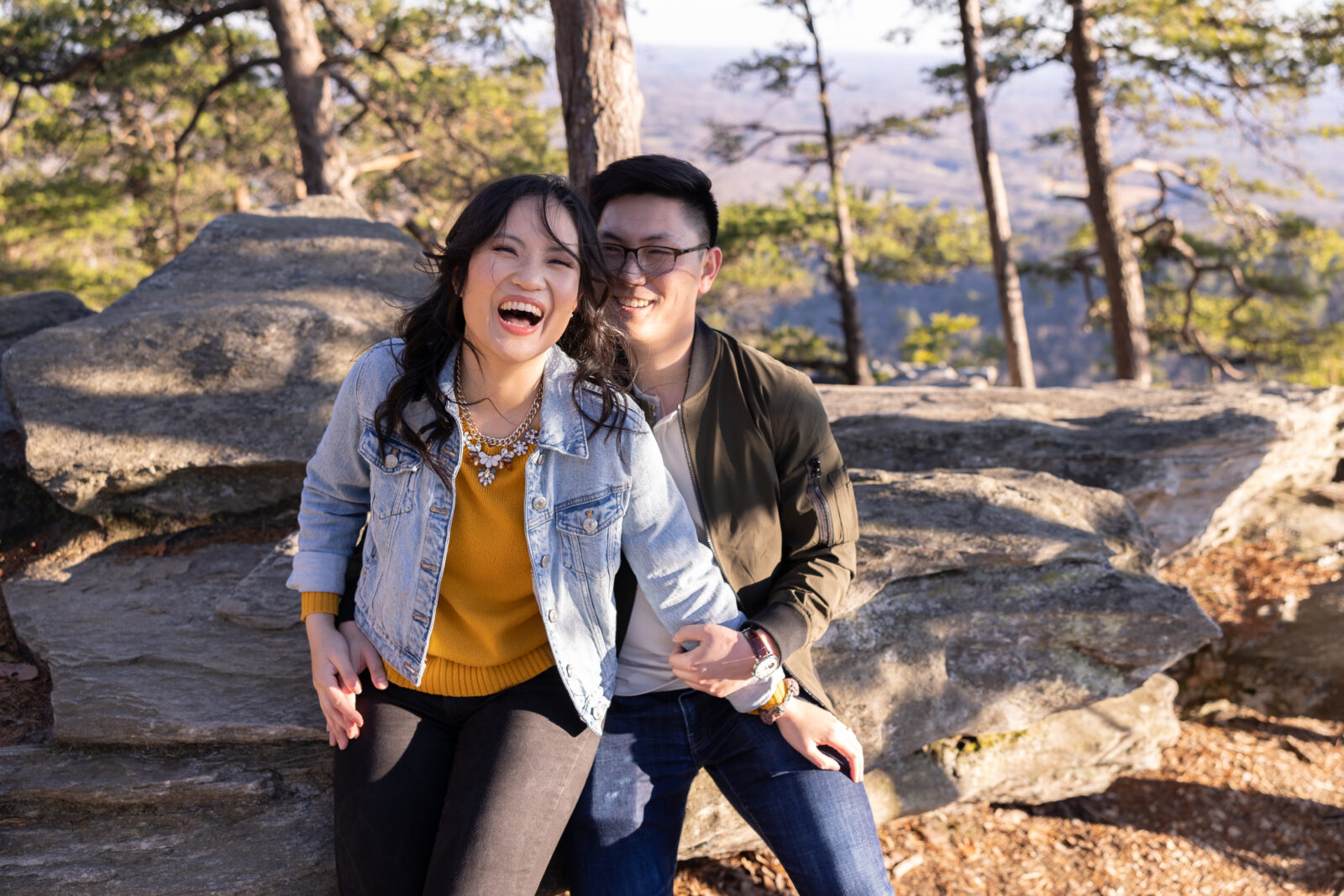 Woman in black jeans and mustard sweater with a jean jacket with a bib statement necklace sits next to man with blue jeans cream shirt and an army green bomber jacket on a bolder at Hanging Rock State Park