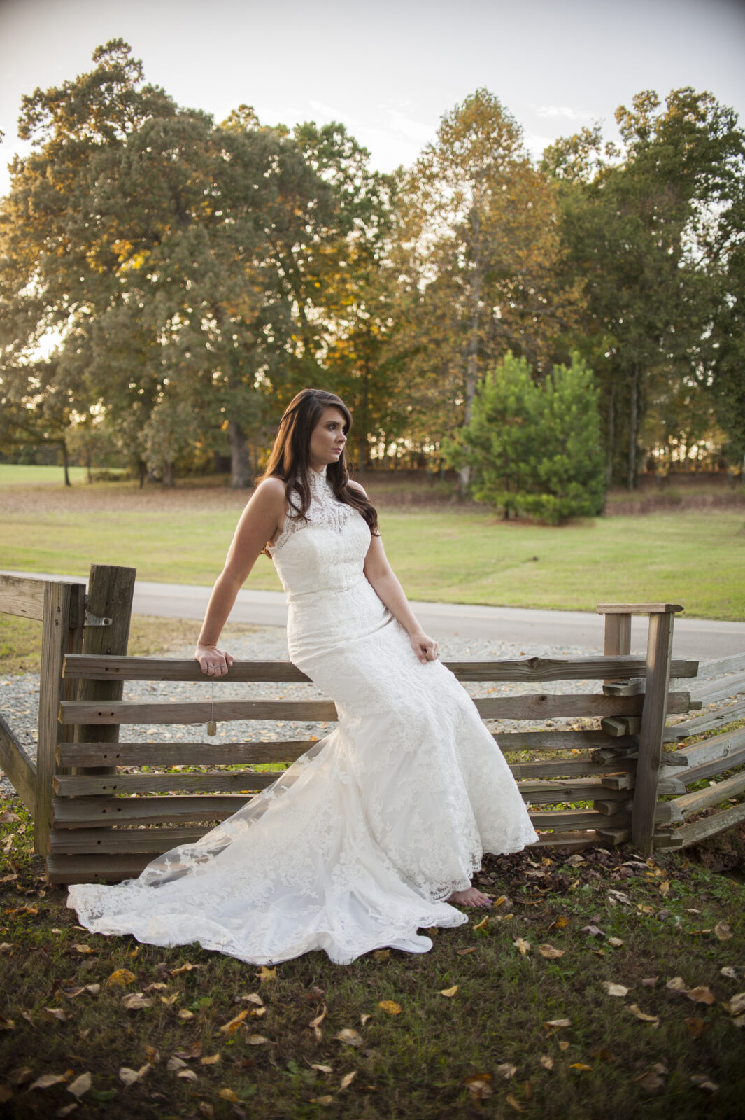 Bride in off white lace detailed halter neckline wedding dress sitting on wood fence at sunset with hair down in soft curls