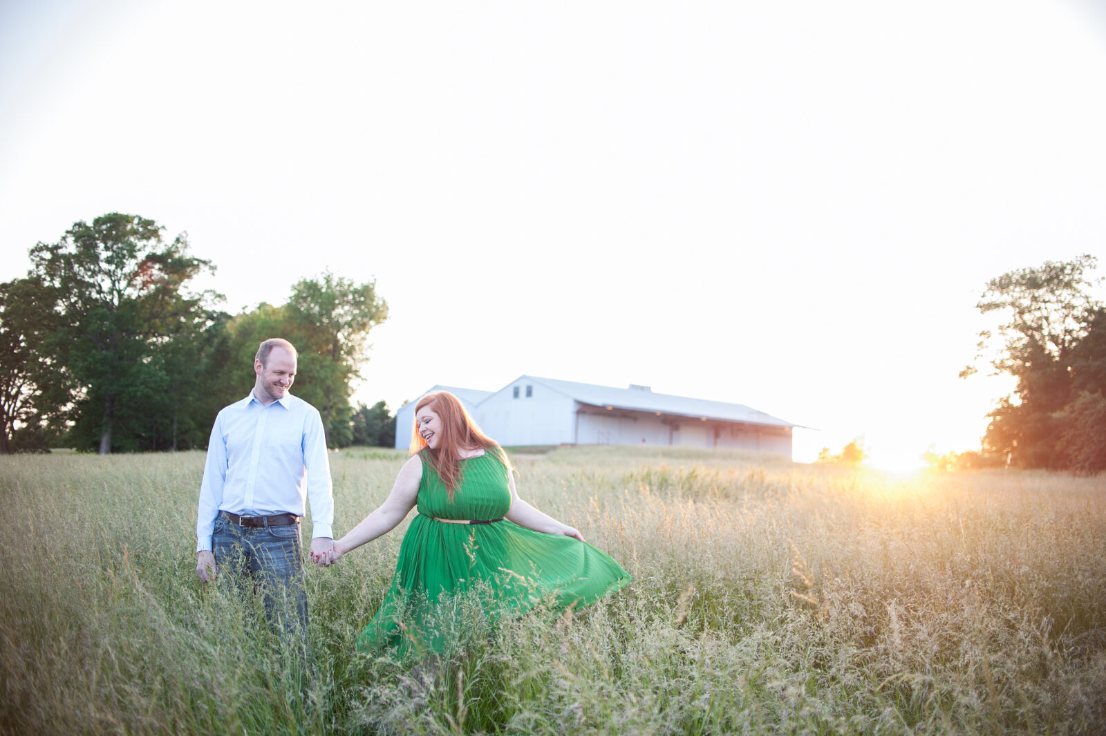 Woman in bright green dress with gold belt and bright red hair twirls in a field of tall grass at North Carolina Museum of Arts holding hands with man who is wearing blue jeans with a brown belt and light blue button down shirt as the sun sets