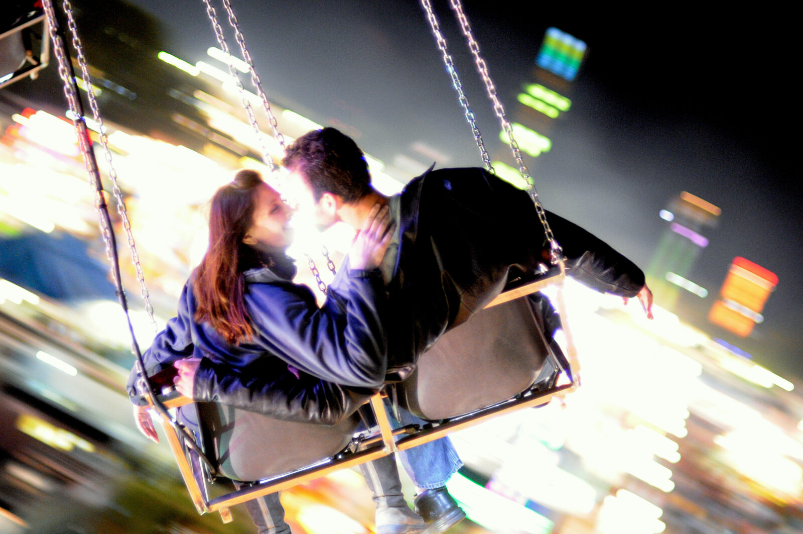 Woman in blue jacket and brown hair embraces a man in a black jacket on suspended swings at the North Carolina State Fair with blurred lights 