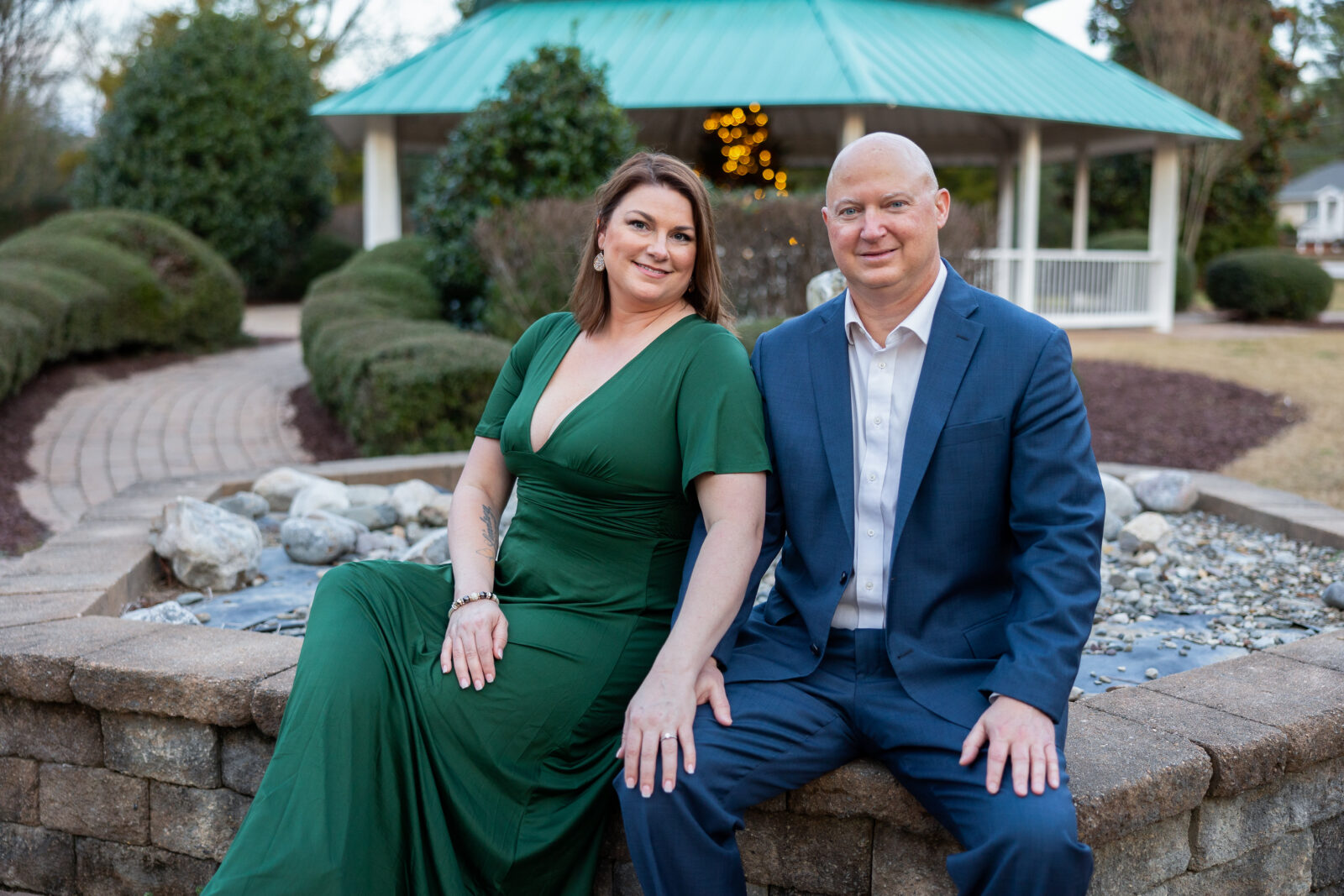 Woman in Green gown with a deep v neckline sitting next to a man with a Blue suit and white button down shirt in front of Rolesville park gazebo with Christmas lights
