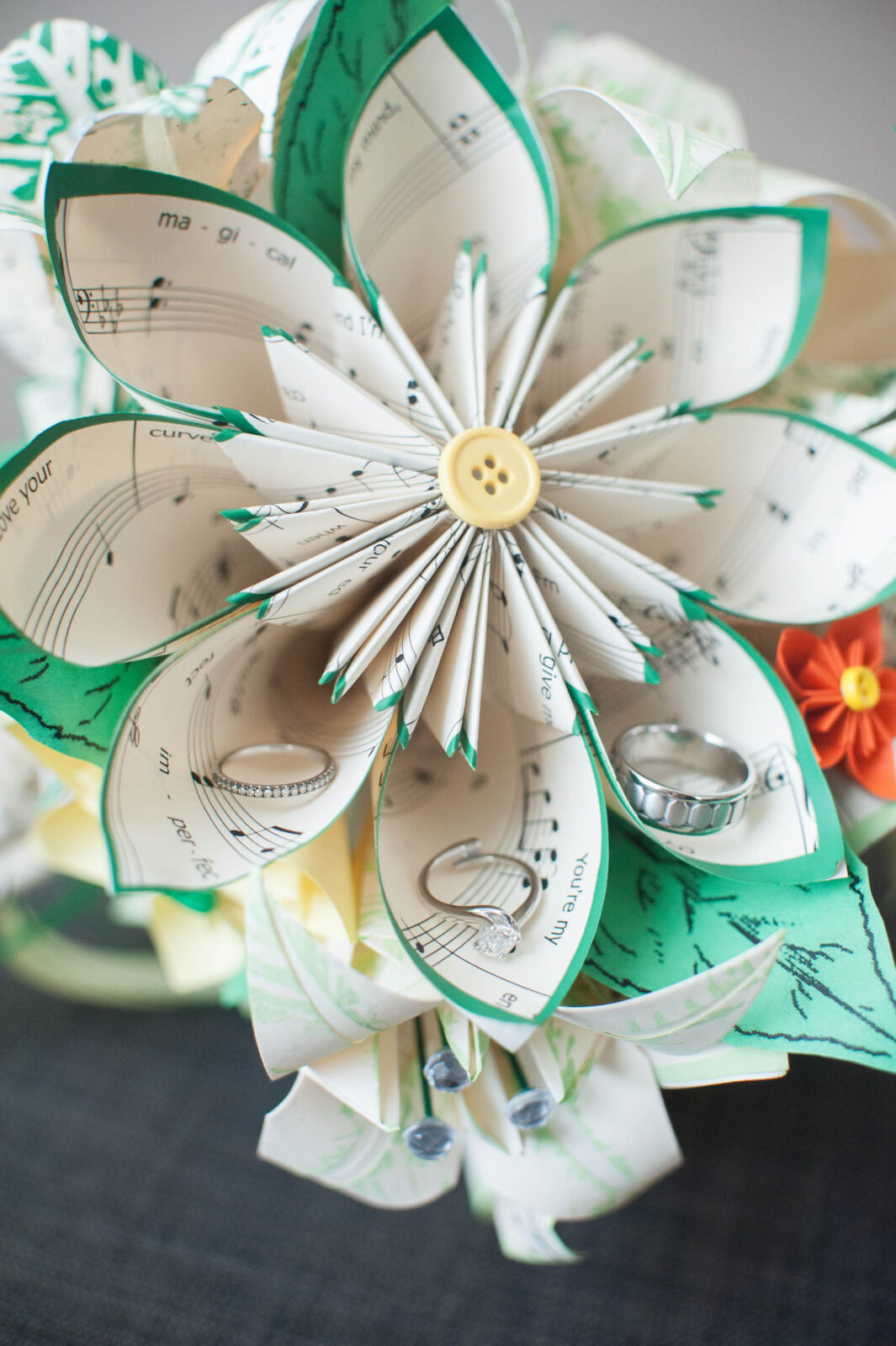 Paper flowers made from sheet music personally picked for each bridesmaid, designed in a green color theme  and a yellow button as center, with rings placed in aspects of that paper flower