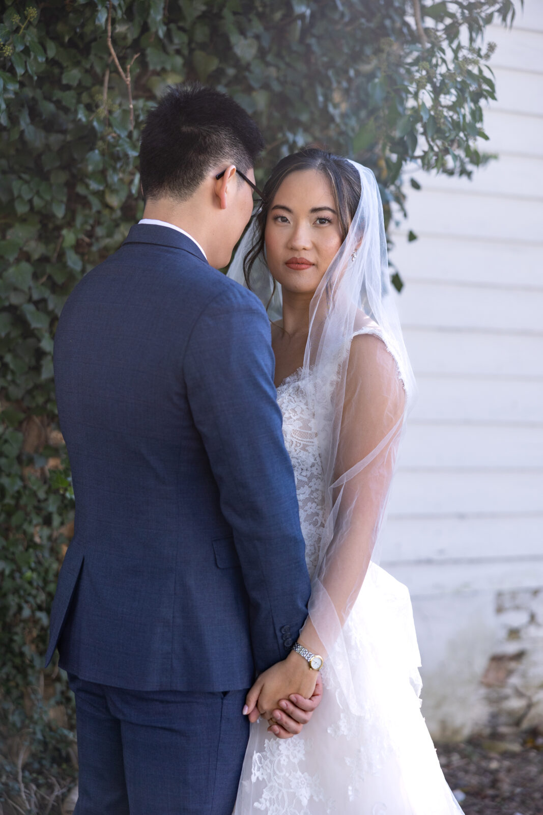 Asian bride in beautiful white lace wedding dress with cathedrial length veil pierces the camera with her gaze as her groom to be in a blue tux holds her hand at tranquility estates in oxford nc