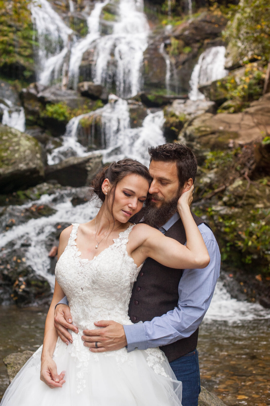 Bride in sweetheart neckline of an offwhite laced detailed dress relaxes into groom wearing a blue button down and vest in front of waterfalls near asheville nc