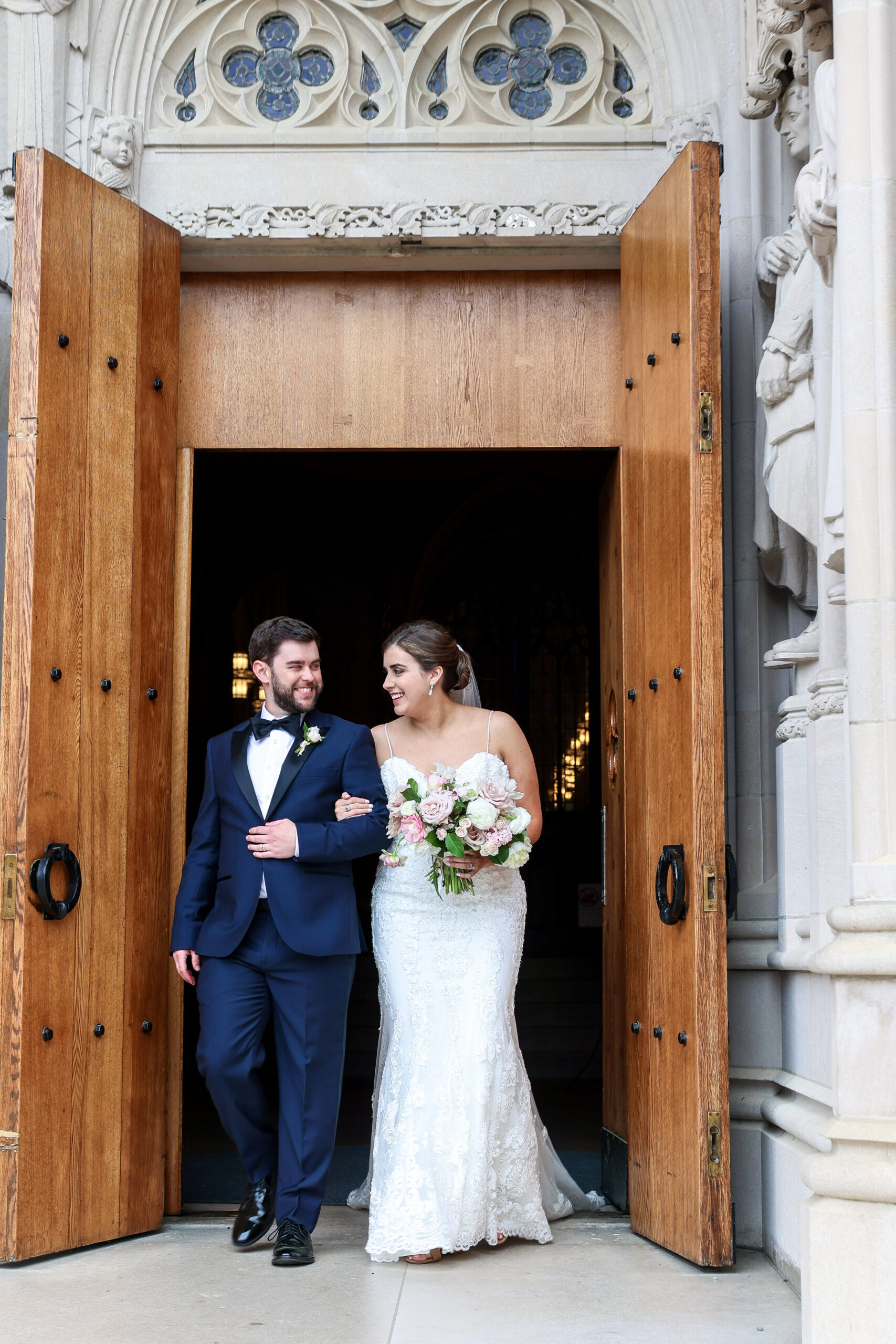 Bride in sweetheart neckline dress smiling and looking at groom with beard and dark brown hair in a blue tux exiting duke chapel