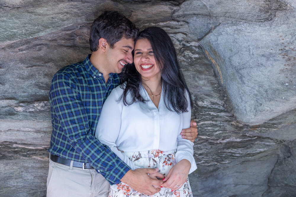 Woman in cream blouse and floral skirt being hugged by man in checkered blue and green button down and khaki pants in front of boulder at hanging rock state park