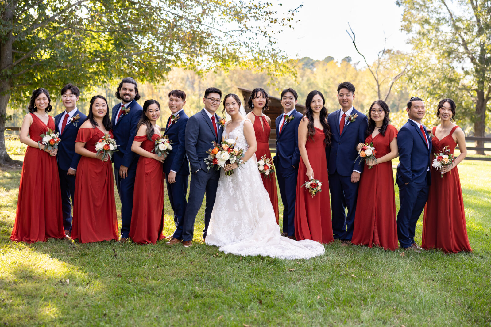 Large bridal party formal with red chiffon bridesmaid dresses and royal blue tuxes for groomsmen on the lawn of an estate.  Bride wearing a lace detailed sweetheart neckline dress with an eclectic bouquet of fall flowers