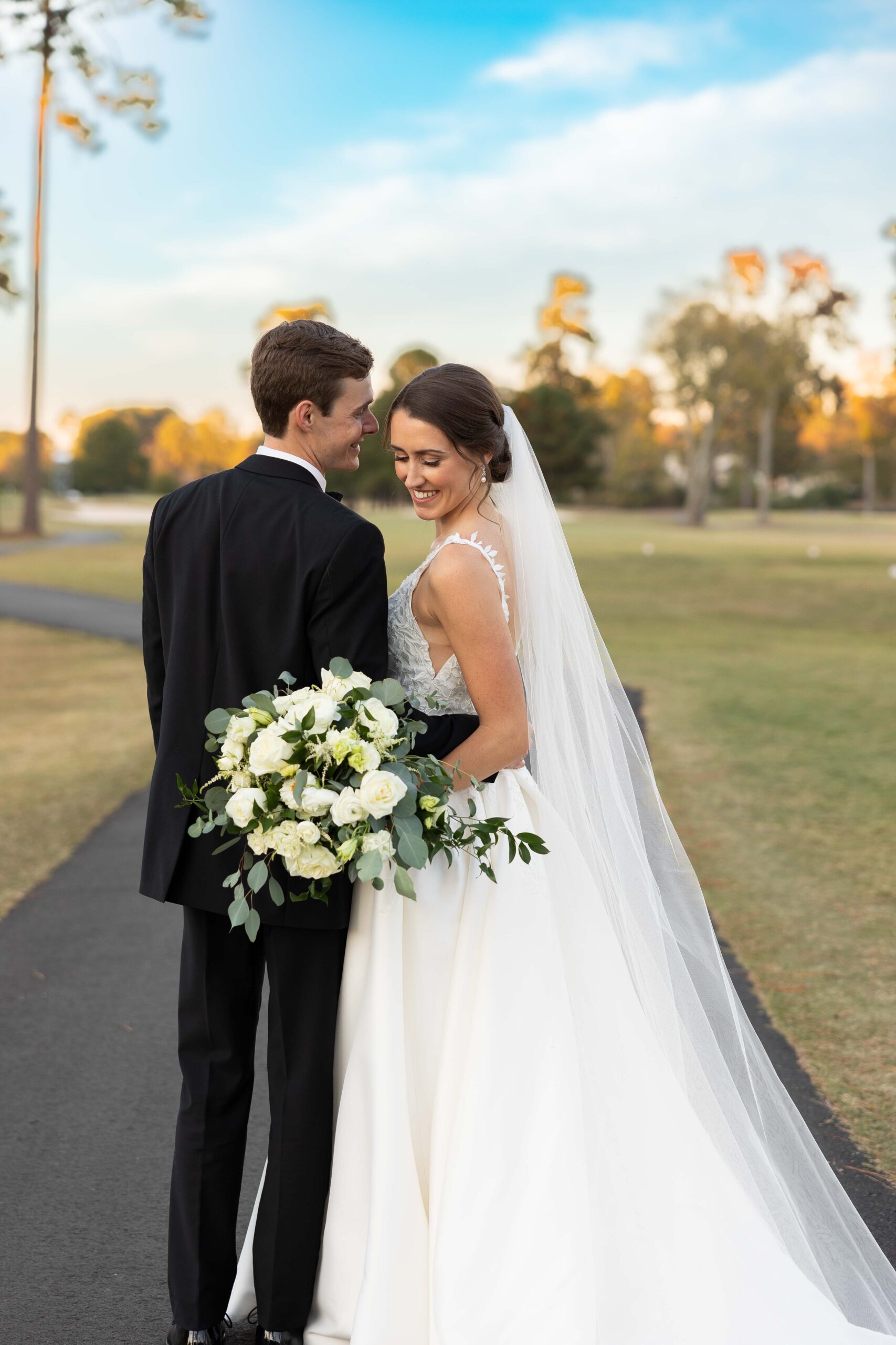 Bride in plunging v lace detailed wedding dress hugs groom in black tux holding. alarg bouquet of white flowers at prestonwood country club