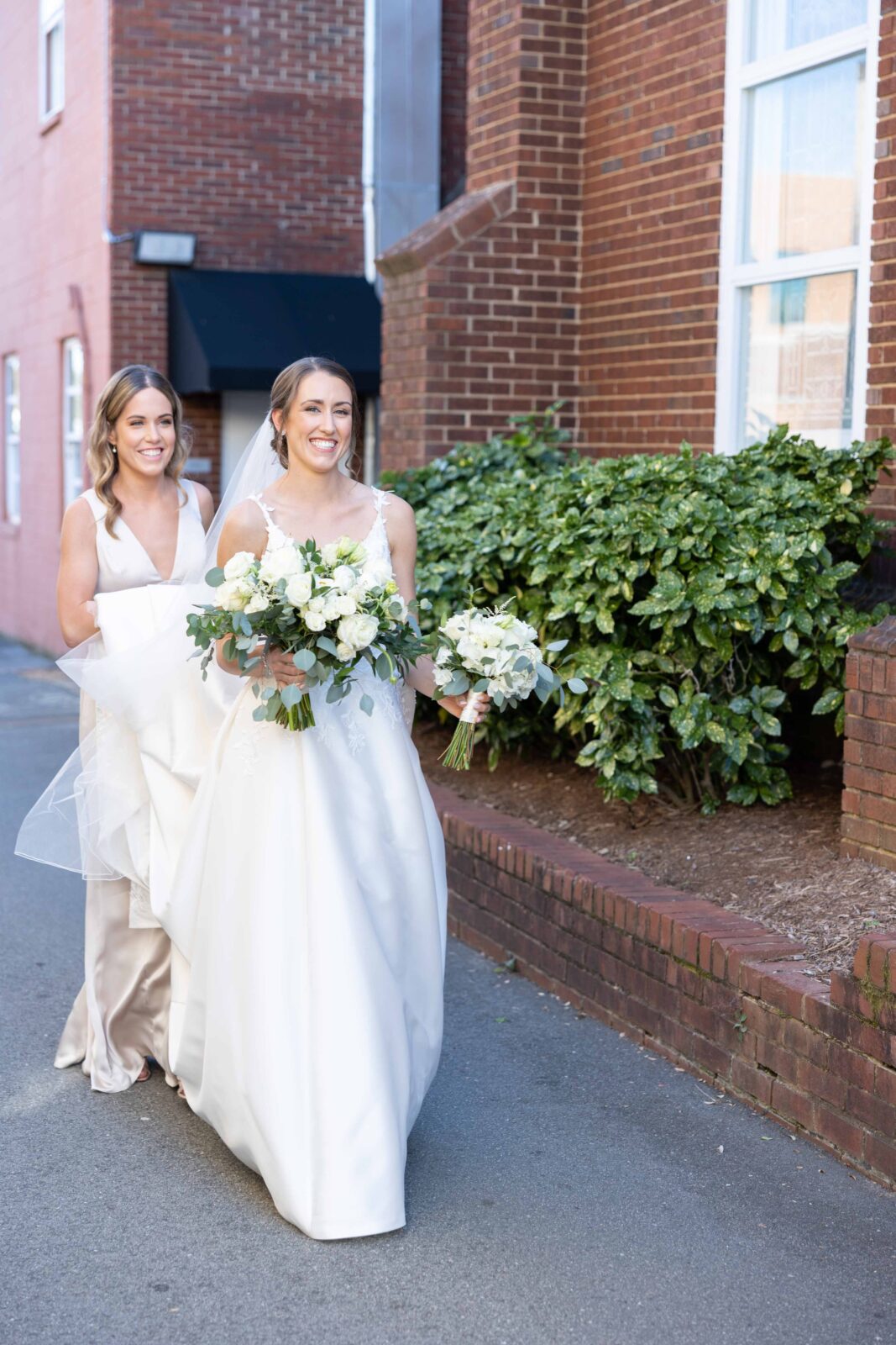 Bride wearing plunging v necked wedding dress with lace floral detail and cathetrial length train held by bridesmaid in gold satin dress