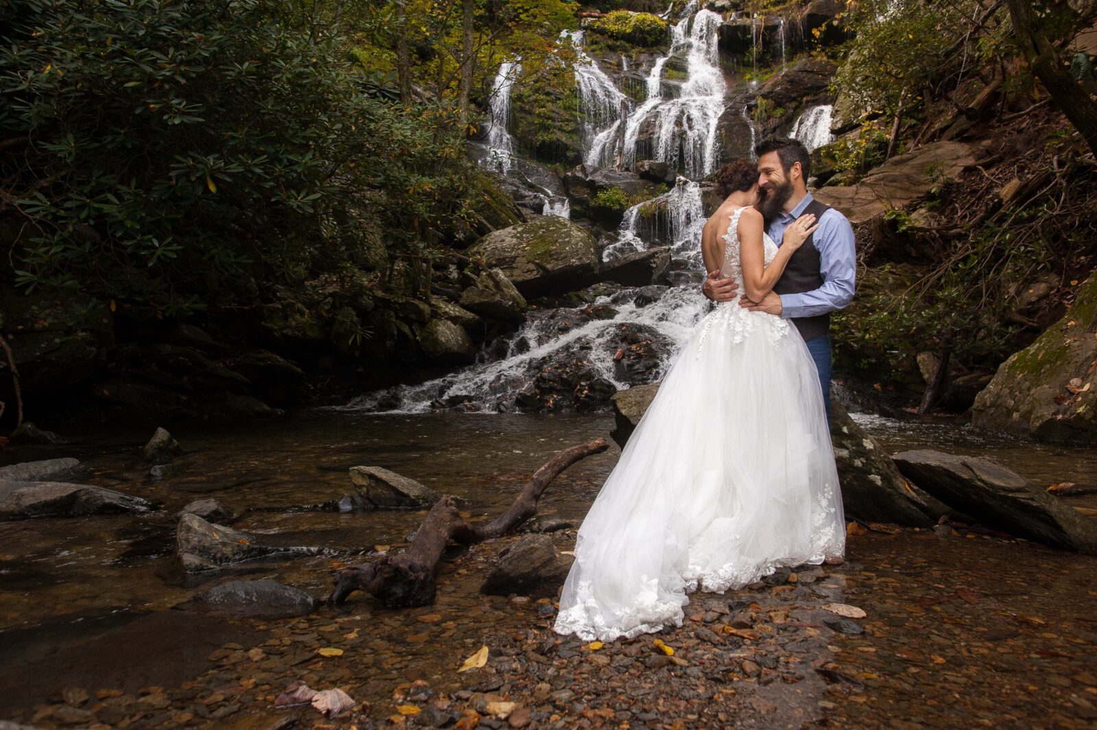 Bride in large lace dress with full skirt and open back dress hugging groom in jeans, blue button down and grey vest in front of a waterfall in asheville nc